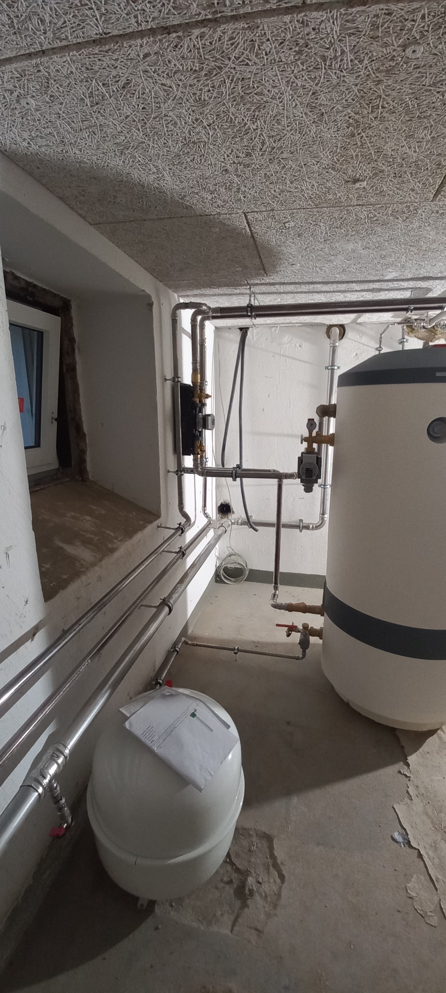 Kocioł grzewczy z osprzętem hydraulicznym, rurami i naczyniem przeponowym w surowym pomieszczeniu z oknem i widocznymi śladami użytkowania na podłodze.