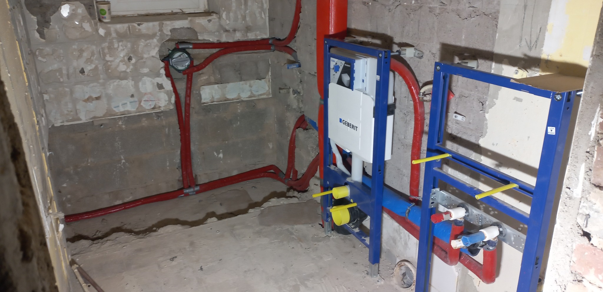 Surowy stan łazienki w trakcie remontu: widoczne instalacje wodne w czerwonych rurach i niebieskie stelaże podtynkowe Geberit, ściany z odsłoniętym murem i resztkami starej farby.
