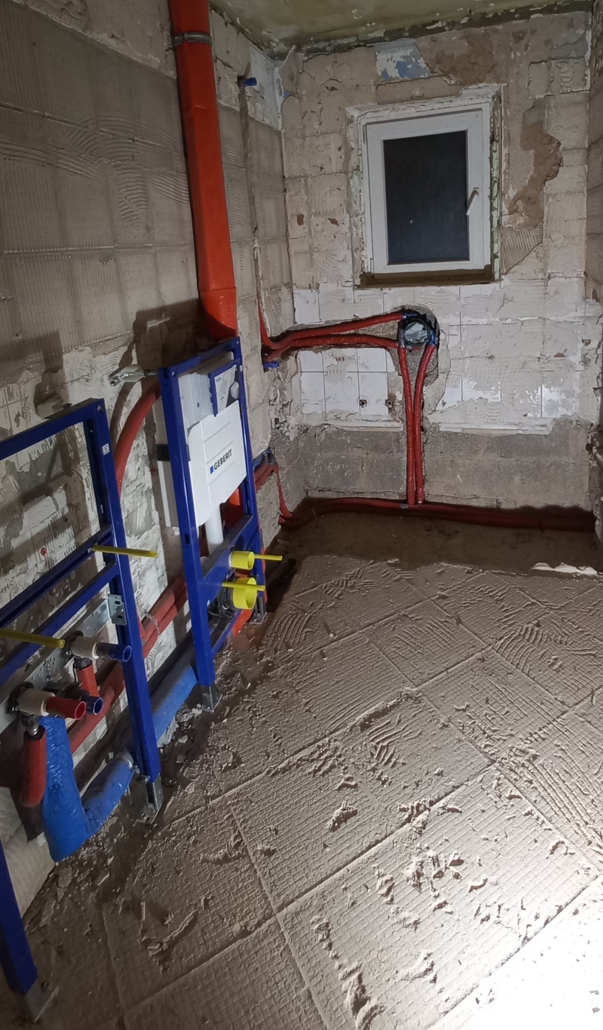 Surowy stan łazienki w trakcie remontu: widoczna instalacja podtynkowa Geberit, rury instalacyjne w kolorze czerwonym i niebieskim, ściany z resztkami płytek i nierównym tynkiem, przygotowana...