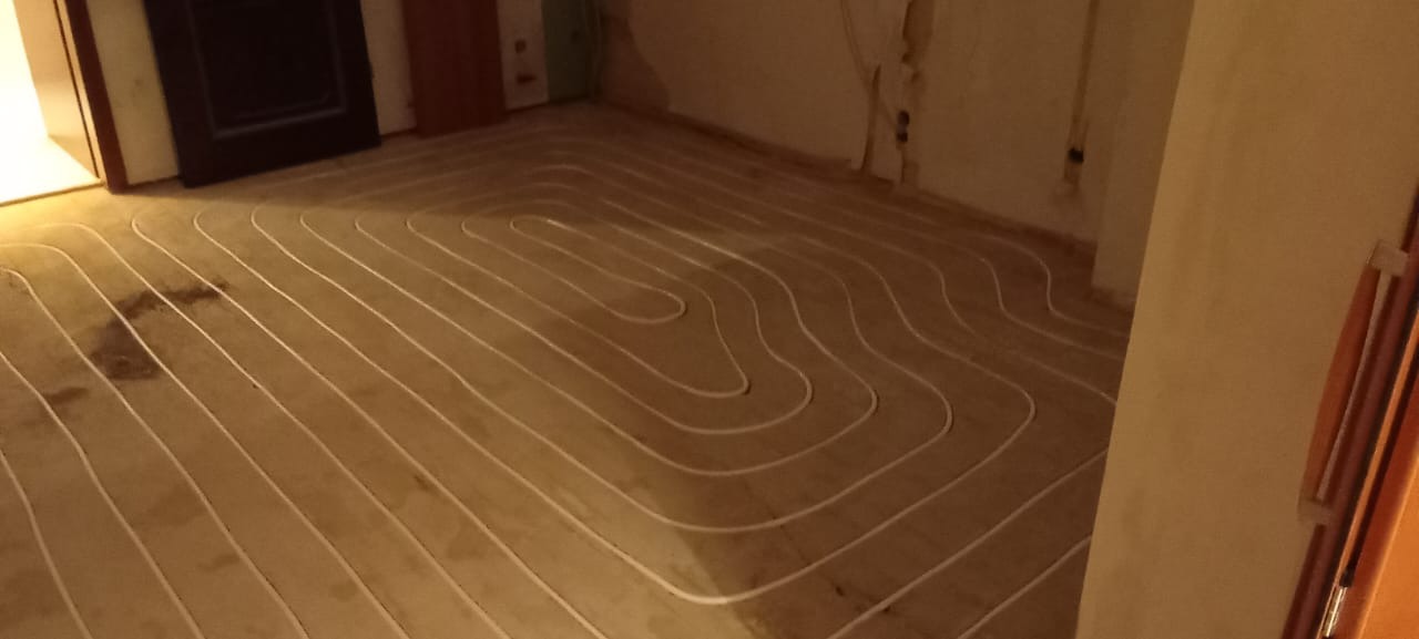 Białe rurki systemu grzewczego ułożone w pętle na drewnianej podłodze w trakcie montażu, widoczne plamy zabrudzeń na podłodze.