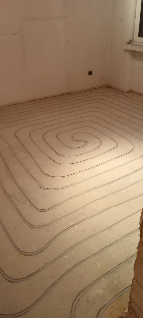 Rury spiralnie ułożone w wylewce podłogowej, przygotowane do montażu systemu grzewczego w jasnym pomieszczeniu z surowymi ścianami.