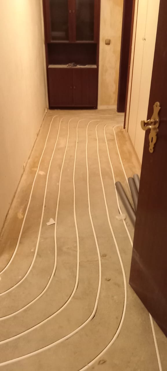 Białe rurki systemu ogrzewania podłogowego ułożone na szarej wylewce w korytarzu, widoczne rury przy ścianie i fragment otwartych drzwi z mosiężną klamką.