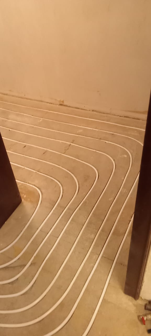 Białe rurki instalacji grzewczej ułożone w pętle na szarym podłożu, widok z góry, fragment ościeżnicy w prawym dolnym rogu.