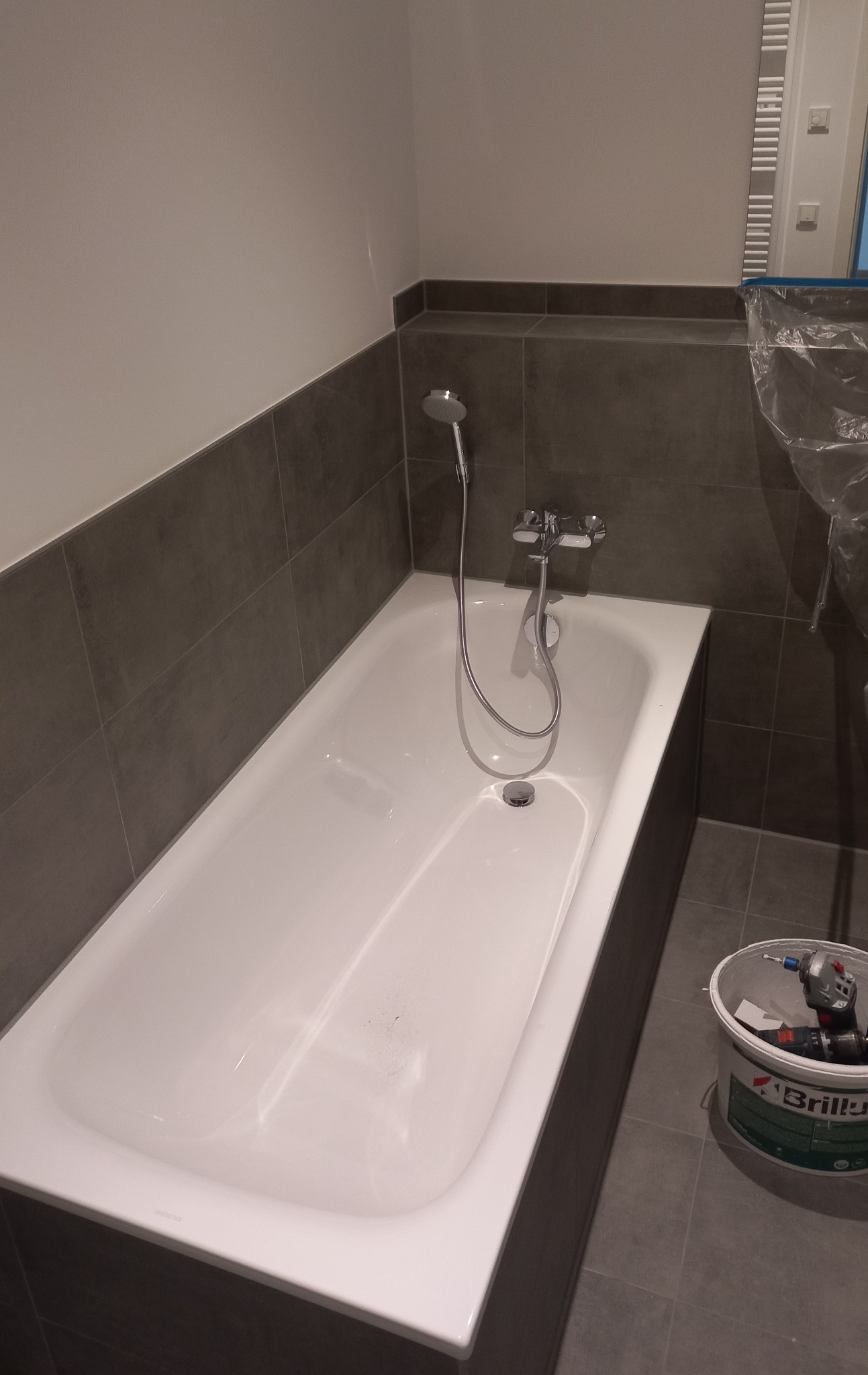 Biała wanna z zamontowaną armaturą, obudowana ciemnymi płytkami, widoczny pojemnik z narzędziami malarskimi i wiertarką.