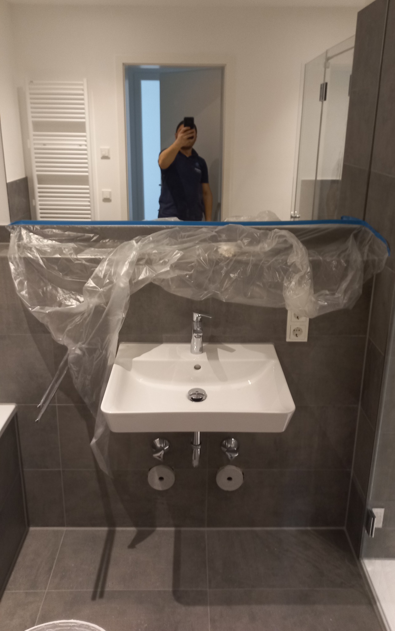 Nowa, biała umywalka zamontowana na szarych płytkach, zabezpieczona folią malarską na czas remontu łazienki, widoczne przyłącza i bateria, odbicie osoby robiącej zdjęcie w lustrze.