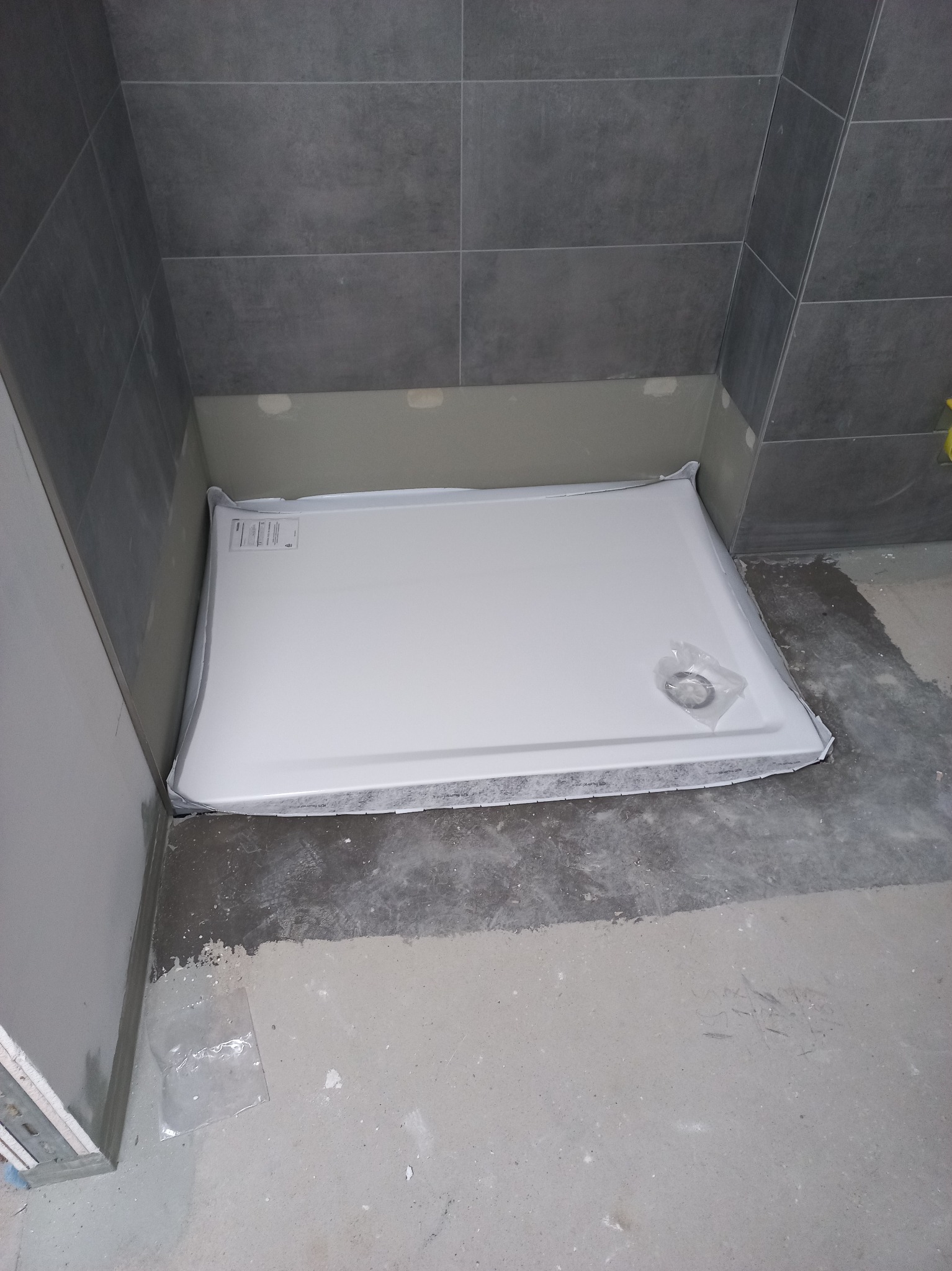 Biały brodzik w folii ochronnej, przygotowany do montażu w narożniku łazienki we Wrocławiu, widoczne szare płytki na ścianie i surowa wylewka na podłodze.