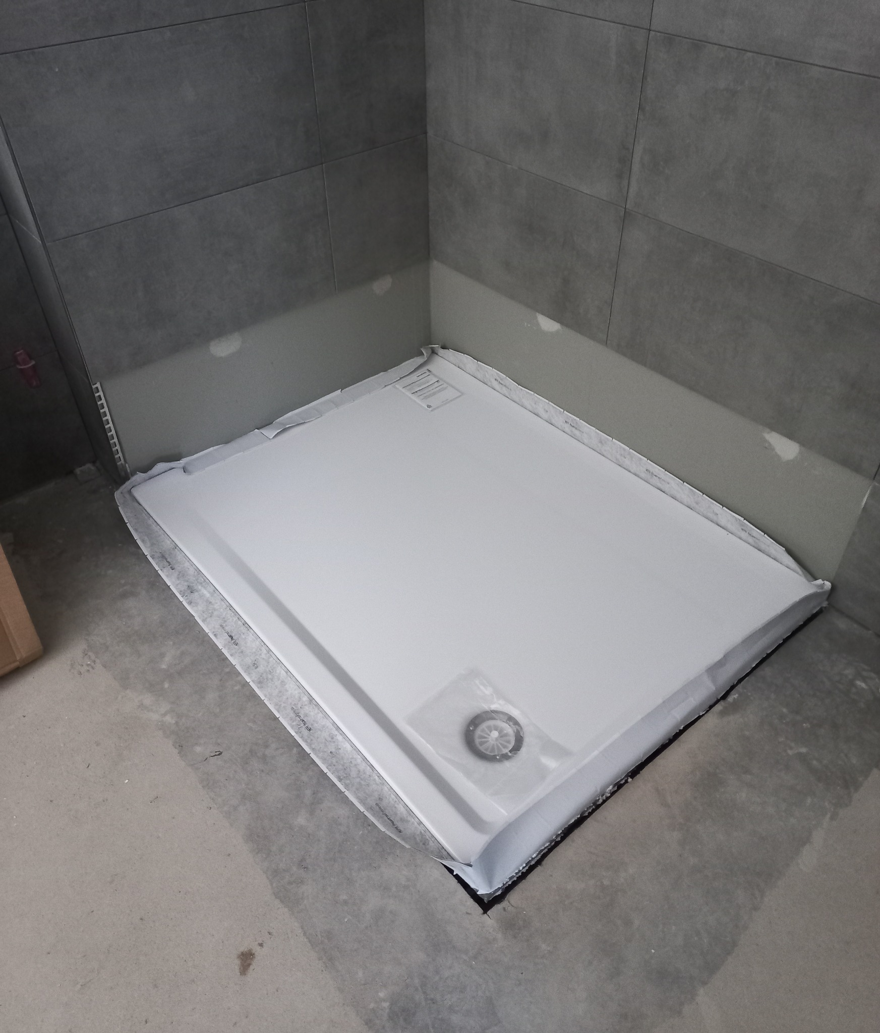 Biały brodzik prysznicowy w folii ochronnej, umieszczony w rogu łazienki z szarymi płytkami, widoczne elementy montażowe w opakowaniu