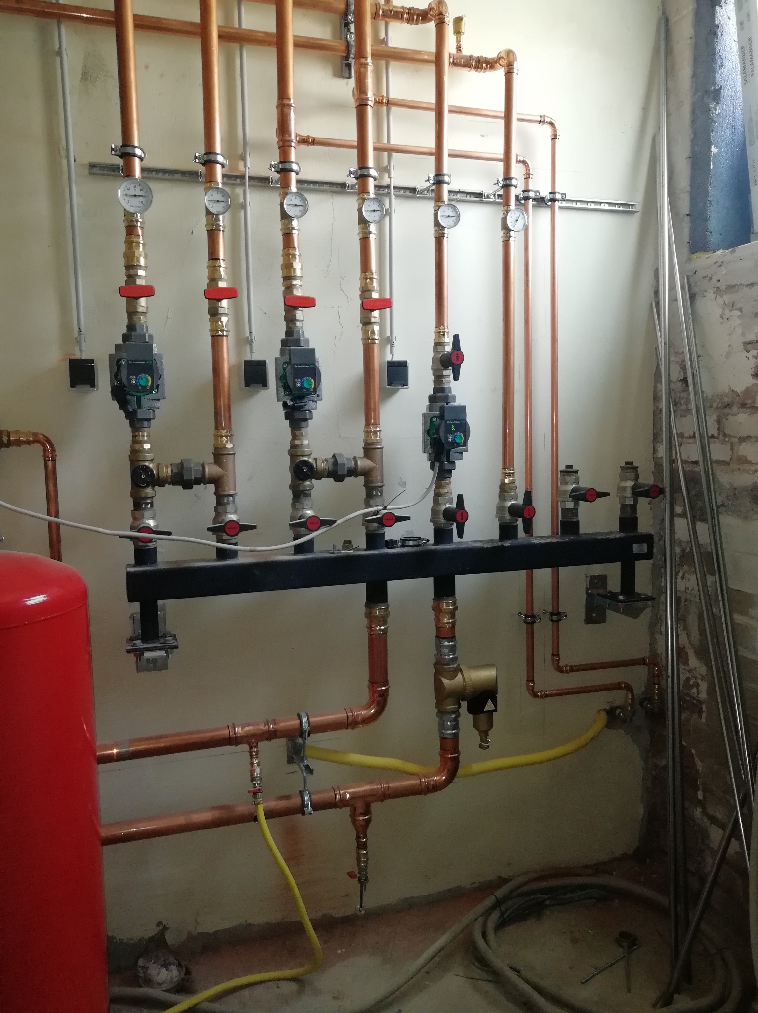 Rozdzielacz ogrzewania podłogowego z miedzianymi rurami, manometrami i zaworami termostatycznymi na tle surowej ściany w trakcie montażu.