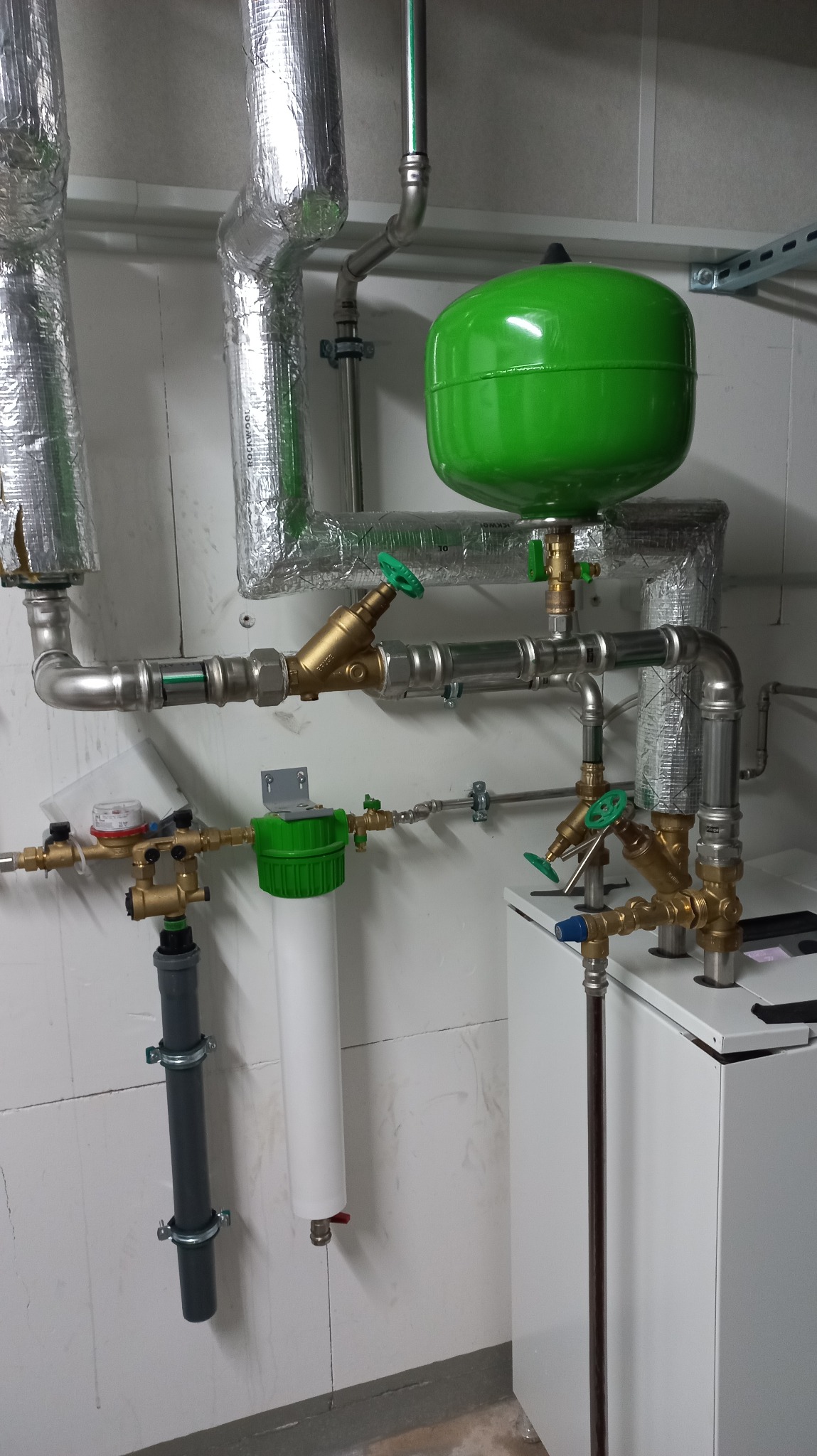 Instalacja hydrauliczna z zielonym zbiornikiem wyrównawczym, filtrem wody i rurami owiniętymi folią izolacyjną, widoczne zawory z zielonymi pokrętłami.