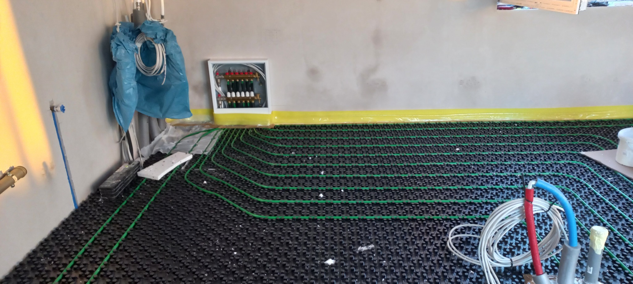 Rozłożona instalacja ogrzewania podłogowego z zielonymi rurkami na czarnej macie, widoczny rozdzielacz w białej obudowie na ścianie, zabezpieczonej żółtą folią, oraz narzędzia i materiały budowlane.