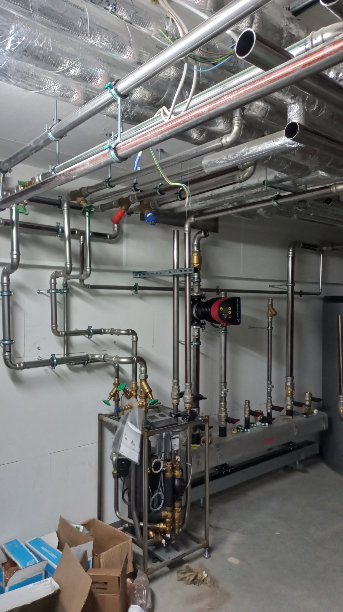 Rozbudowana instalacja grzewcza z licznymi rurami, zaworami i urządzeniami, zamontowana pod sufitem z izolacją, widoczne elementy kotłowni w surowym pomieszczeniu.