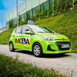 Daniel Smoter - Jasnozielony samochód nauki jazdy Imola z symbolem 'L' na dachu, zaparkowany na szutrowej drodze, z zielonym ekranem akustycznym porośniętym bluszczem w tle.