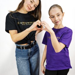 Daniel Smoter - Portret dwóch uśmiechniętych kobiet, dorosłej i nastolatki, ustawionych blisko siebie na białym tle, obie mają na sobie koszulki z logo firmy i formują dłońmi kształt serca.