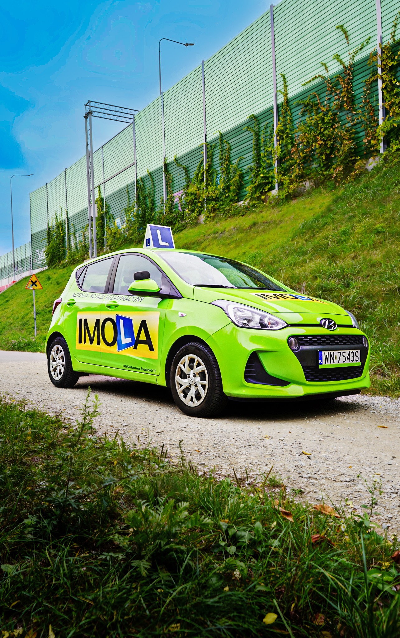 Jasnozielony samochód nauki jazdy Imola z symbolem 'L' na dachu, zaparkowany na szutrowej drodze, z zielonym ekranem akustycznym porośniętym bluszczem w tle.