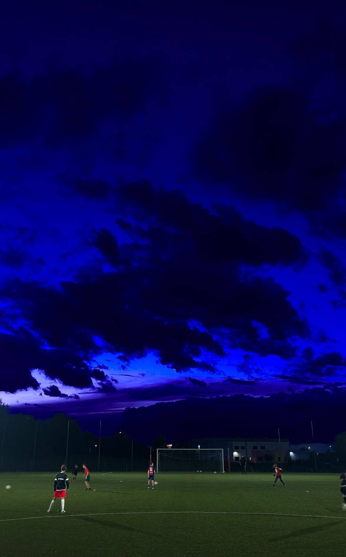Wieczorny trening piłkarski na oświetlonym boisku, ciemne chmury na intensywnie niebieskim niebie.