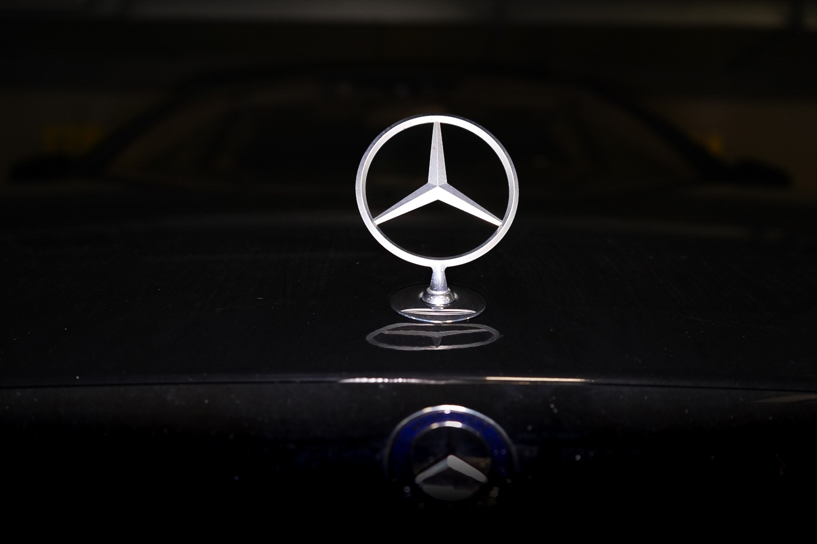 Chromowany znaczek Mercedes-Benz na czarnej masce samochodu, widoczny w słabym oświetleniu z odblaskiem na lakierze.