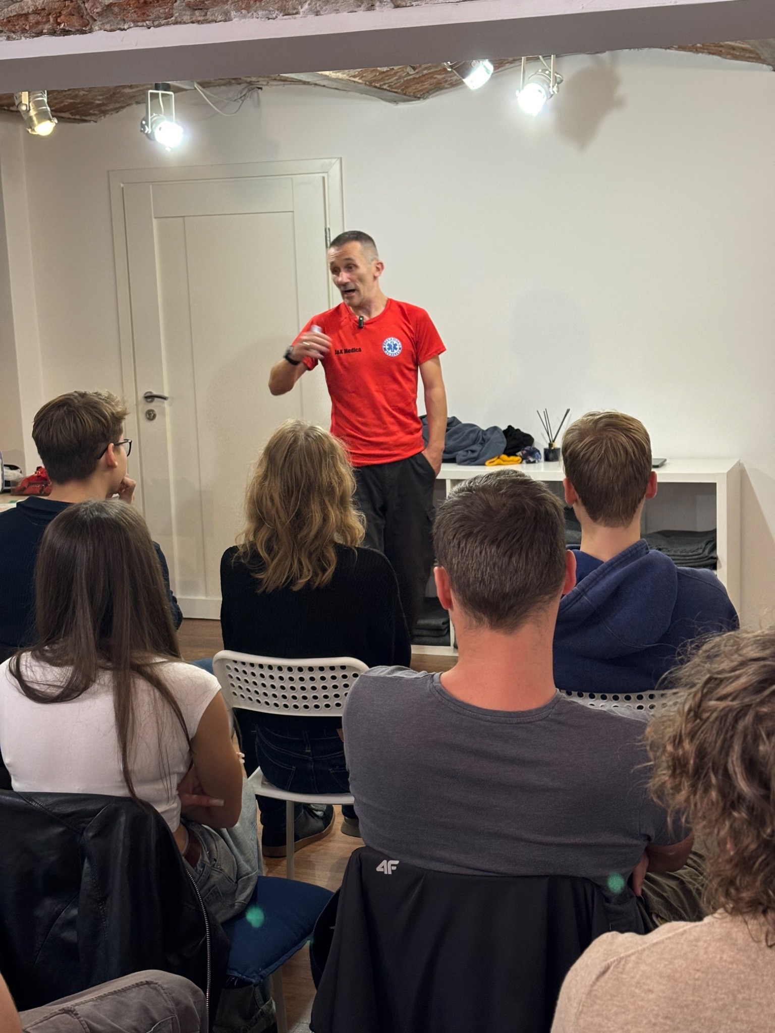 Instruktor w czerwonej koszulce z logo medycznym prowadzi szkolenie z pierwszej pomocy dla grupy słuchaczy w pomieszczeniu z ceglanym sufitem.
