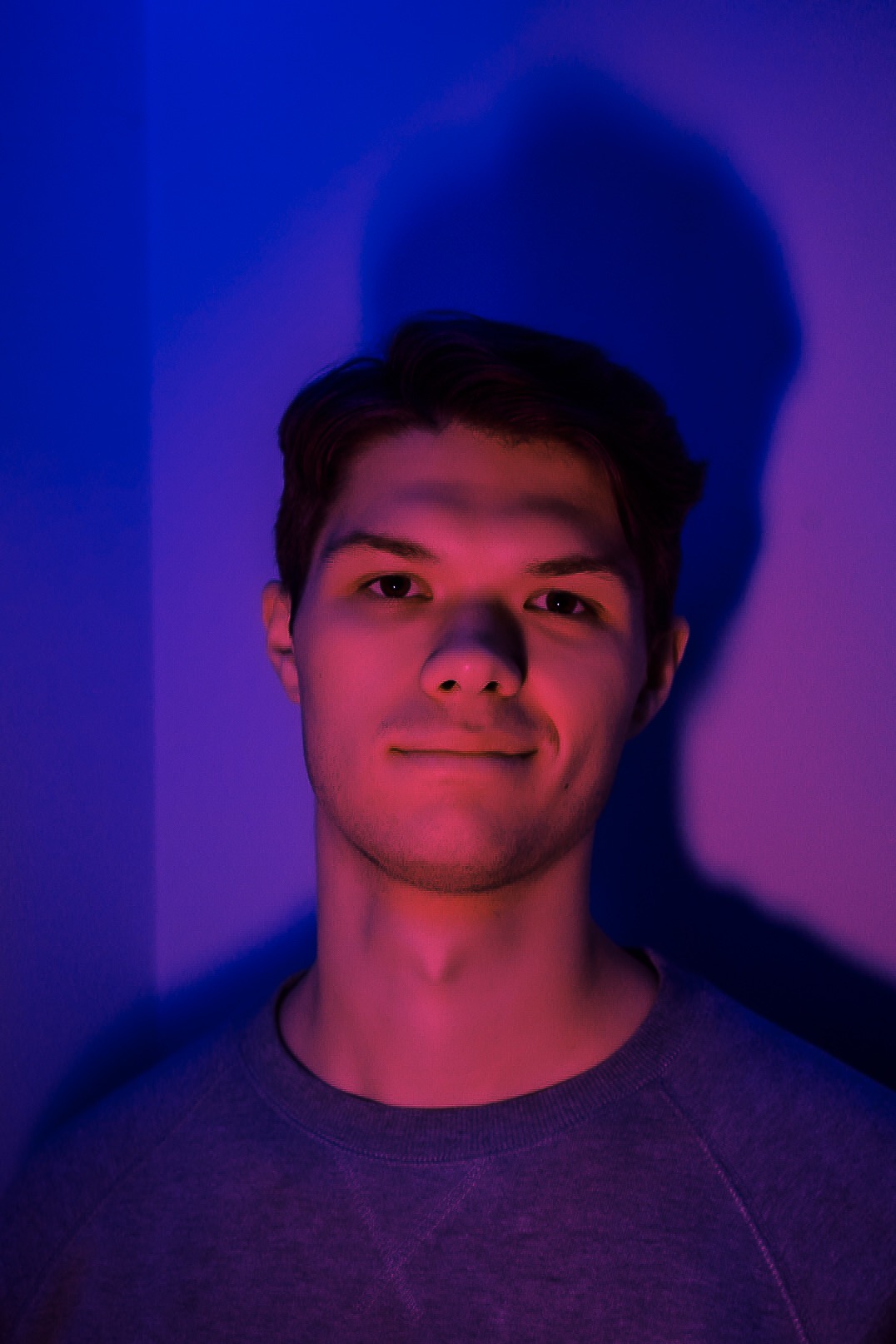 Portret młodego mężczyzny oświetlonego czerwonym i niebieskim światłem, z cieniem na ścianie za nim.