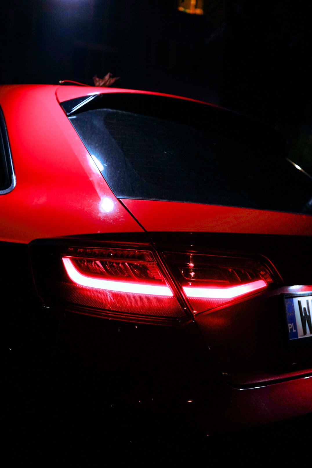 Czerwony samochód z włączonymi tylnymi światłami, widok z tyłu w nocy, widoczna tablica rejestracyjna.
