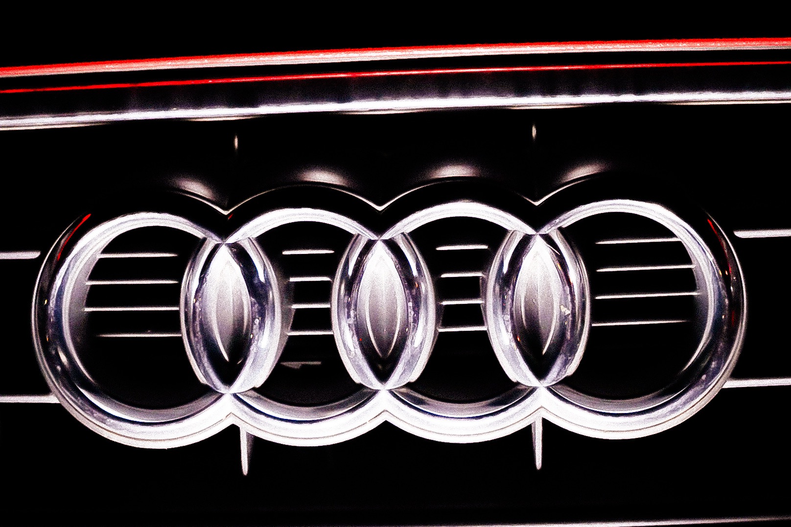 Chromowane logo Audi na czarnym tle grilla samochodowego z subtelnym czerwonym akcentem na górze.