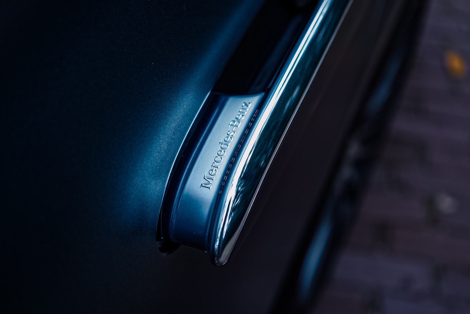 Błyszczący, chromowany napis 'Mercedes-Benz' na ciemnoniebieskim, matowym lakierze samochodu, uchwycony pod kątem z bliska.
