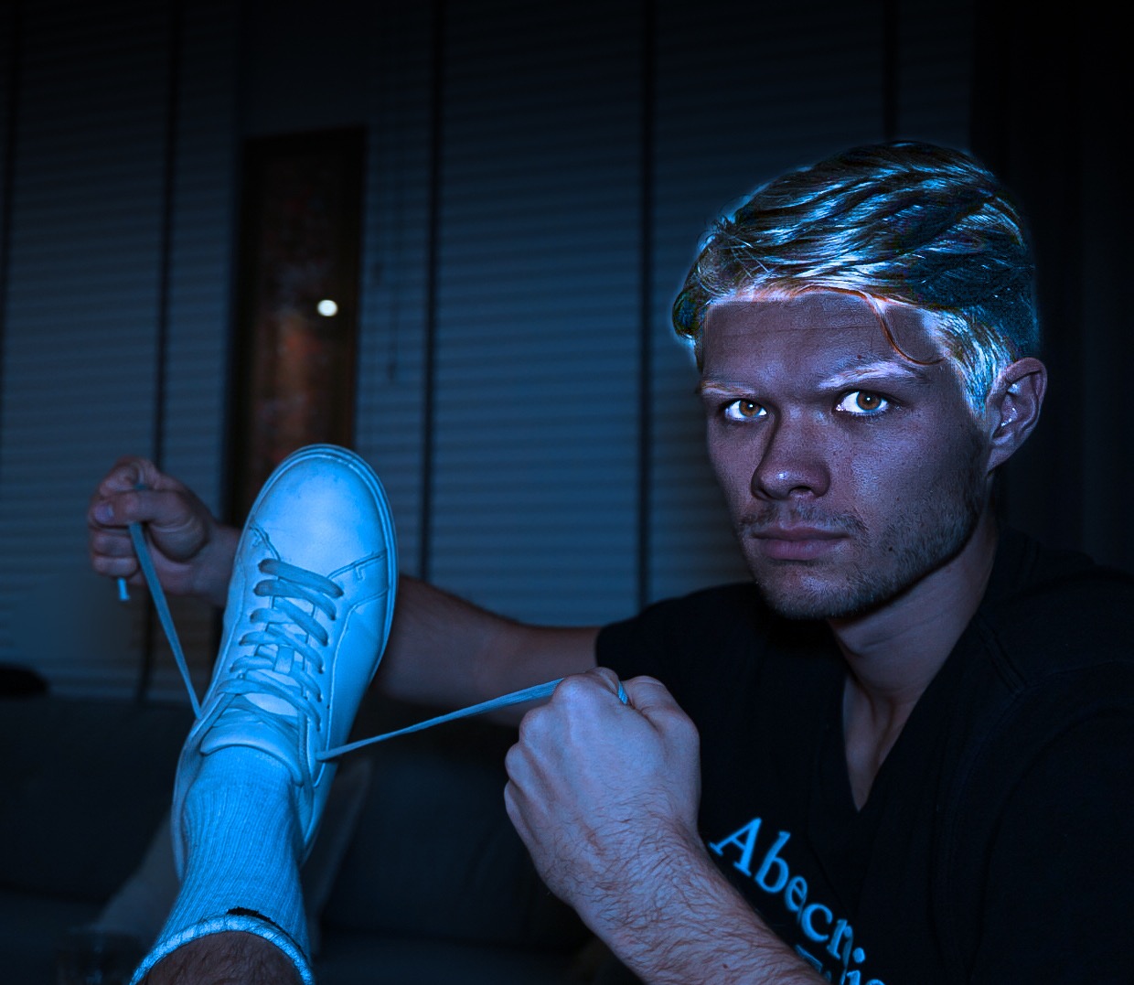 Młody mężczyzna w czarnej koszulce wiąże sznurówki białego tenisówki w chłodnym, niebieskim oświetleniu.