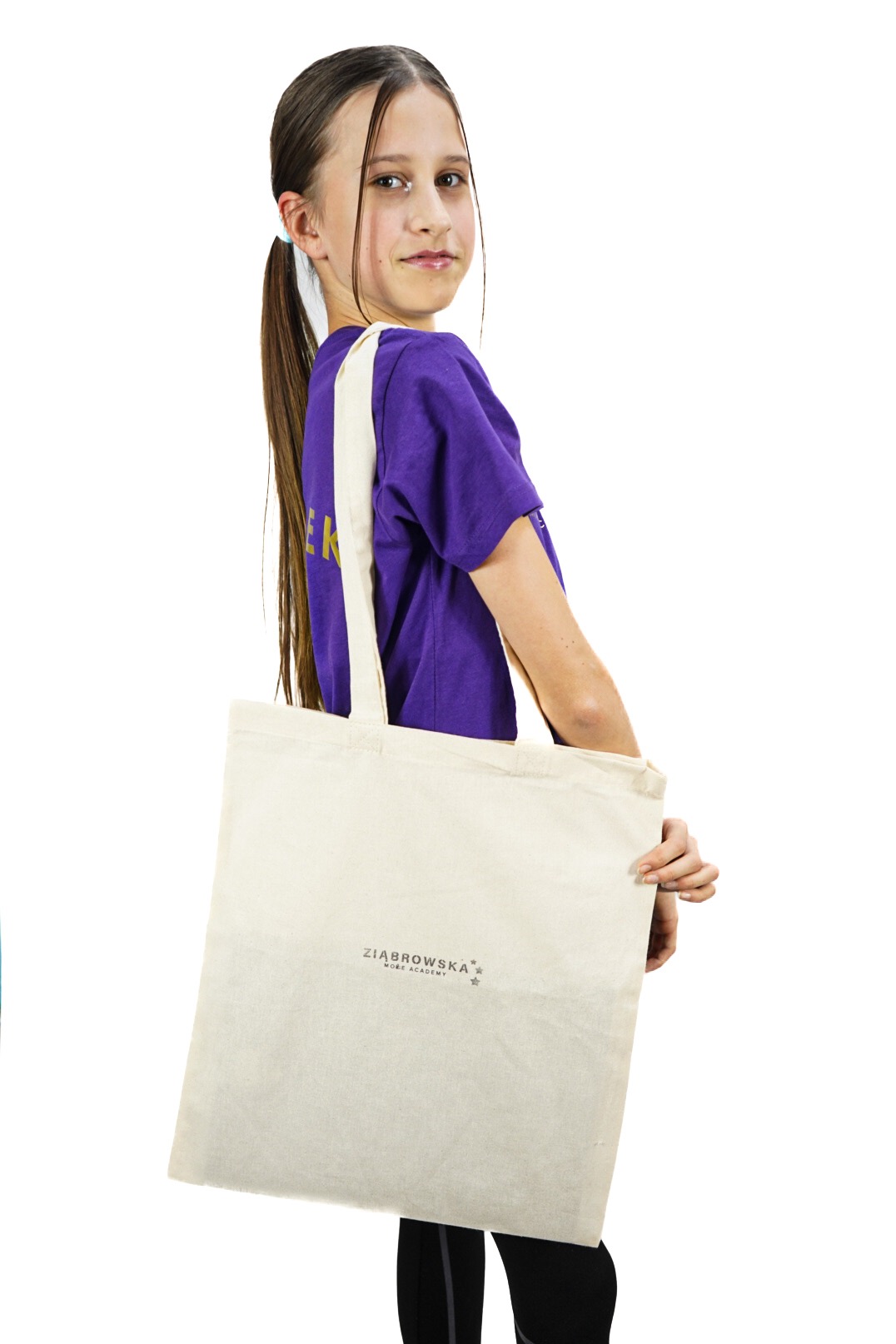 Dziewczynka z długimi włosami w kucykach, ubrana w fioletową koszulkę, trzyma beżową torbę bawełnianą z nadrukowanym logo firmy ZIABROWSKA.