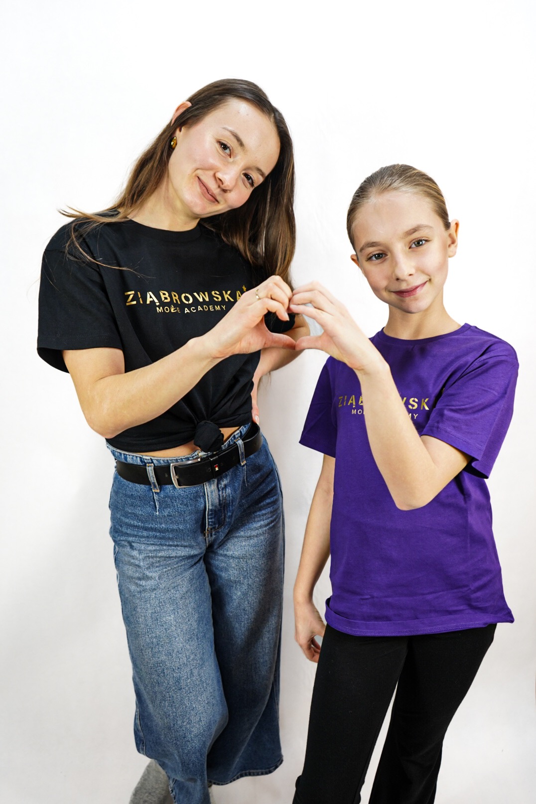 Portret dwóch uśmiechniętych kobiet, dorosłej i nastolatki, ustawionych blisko siebie na białym tle, obie mają na sobie koszulki z logo firmy i formują dłońmi kształt serca.