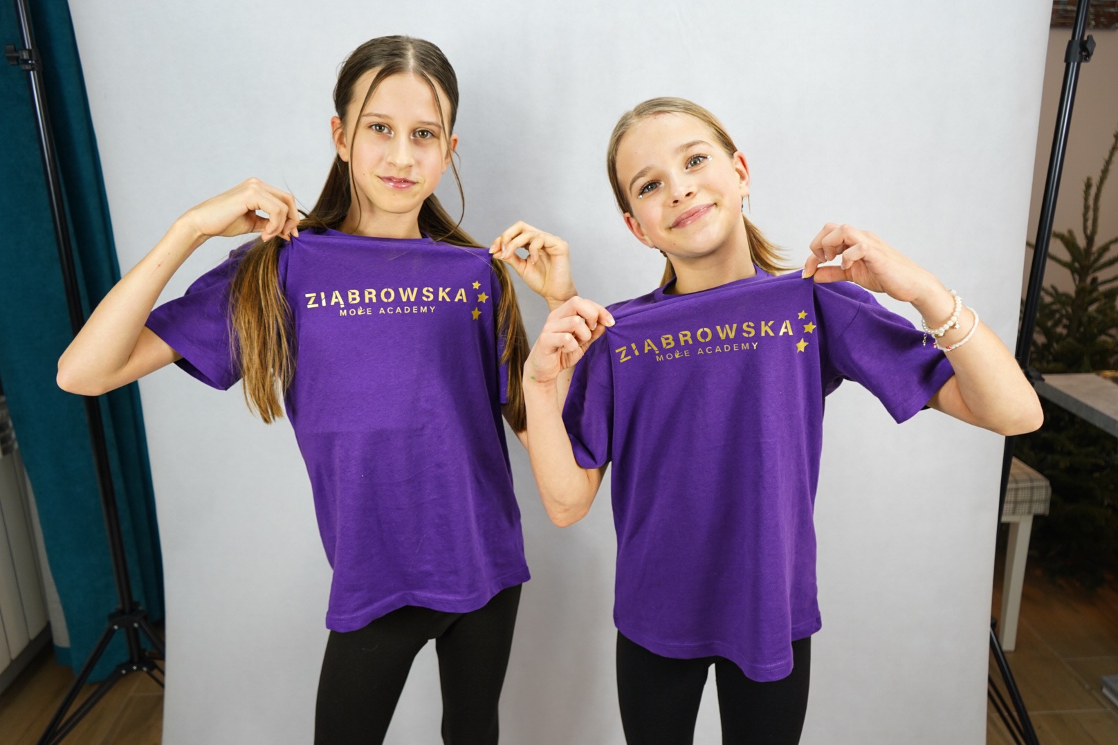Dwie uśmiechnięte dziewczynki w fioletowych koszulkach z logo 'Żiąbrowska Może Academy' na tle studyjnym.