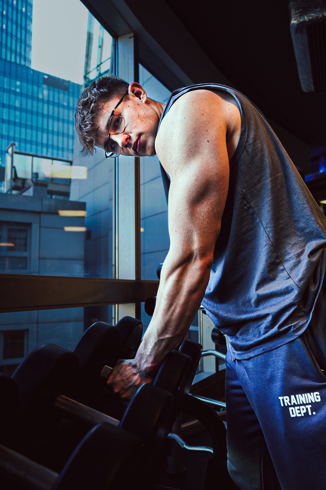 Mężczyzna w okularach, w sportowym stroju, sięga po hantle w siłowni na tle przeszklenia z widokiem na miasto.