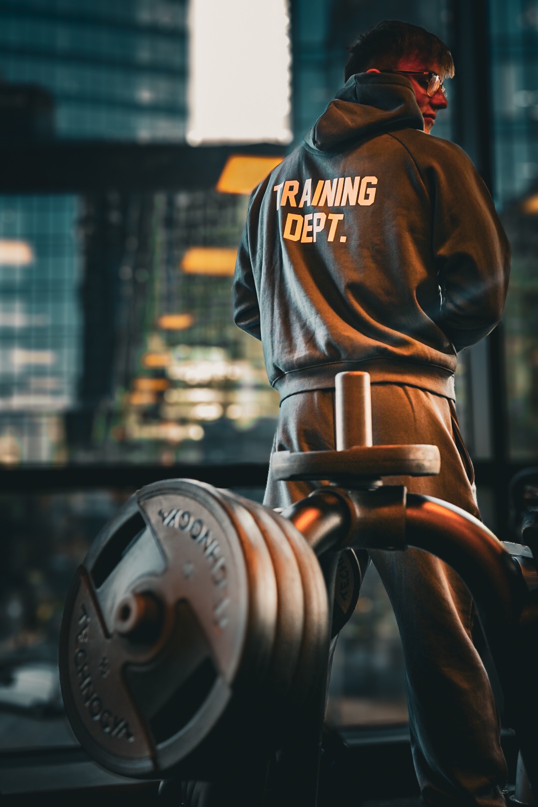 Mężczyzna w dresie z napisem 'TRAINING DEPT.' na plecach stoi obok sztangi w siłowni na tle okna z widokiem na miasto.