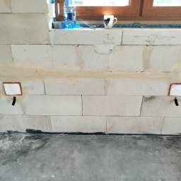 AD MIKOMAX Sp.z o.o. - Ściana z betonu komórkowego z przygotowanymi otworami i rurami do instalacji elektrycznej, widoczne oznaczenia pomiarowe i notatki na ścianie, butelka wody i kubek na parapecie.