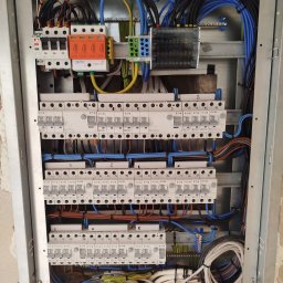 AD MIKOMAX Sp.z o.o. - Rozdzielnica elektryczna w metalowej obudowie z widocznymi bezpiecznikami, przewodami w kolorach niebieskim, czarnym, brązowym i żółto-zielonym oraz zabezpieczeniami przeciwprzepięciowymi SIMTEC...