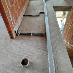 AD MIKOMAX Sp.z o.o. - Wewnętrzna instalacja elektryczna w trakcie montażu: kable w peszlach mocowane do ściany z cegły, pionowa metalowa listwa maskująca, widoczny otwór w ścianie. Surowy stan wykończenia wnętrza.