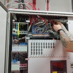 AD MIKOMAX Sp.z o.o. - Otwarta szafa sterownicza z licznymi przewodami i elementami elektrycznymi, ręce elektryka z tatuażem podłączają czerwone kable w górnej części panelu.