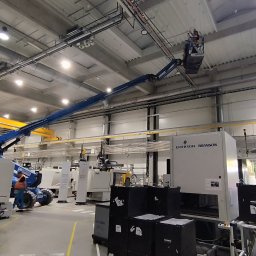 AD MIKOMAX Sp.z o.o. - Pracownik na podnośniku koszowym wykonuje prace elektryczne przy oświetleniu w hali przemysłowej, widoczne rury i kable pod sufitem oraz maszyny produkcyjne.
