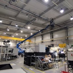 AD MIKOMAX Sp.z o.o. - Pracownik na podnośniku koszowym wykonuje prace na wysokości pod sufitem hali przemysłowej, widoczne rury i przewody pod stropem, w tle maszyny i urządzenia produkcyjne.