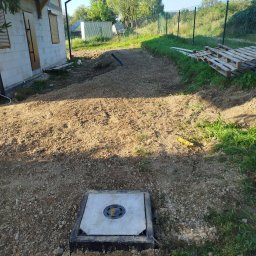 AD MIKOMAX Sp.z o.o. - Studzienka kanalizacyjna z betonową pokrywą osadzona w gruncie, widok na plac budowy domu jednorodzinnego w słoneczny dzień.