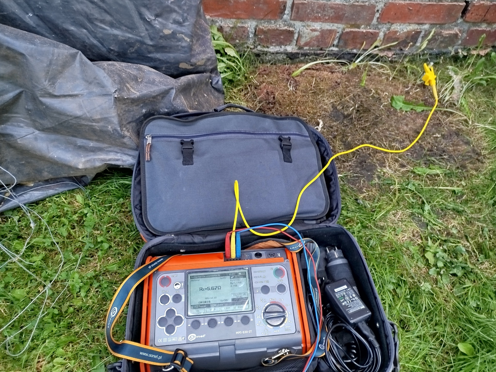 Miernik rezystancji uziemienia Sonel MPI-530-IT w otwartej torbie na trawie, obok ściany z cegły i folii budowlanej. Widoczne przewody pomiarowe.