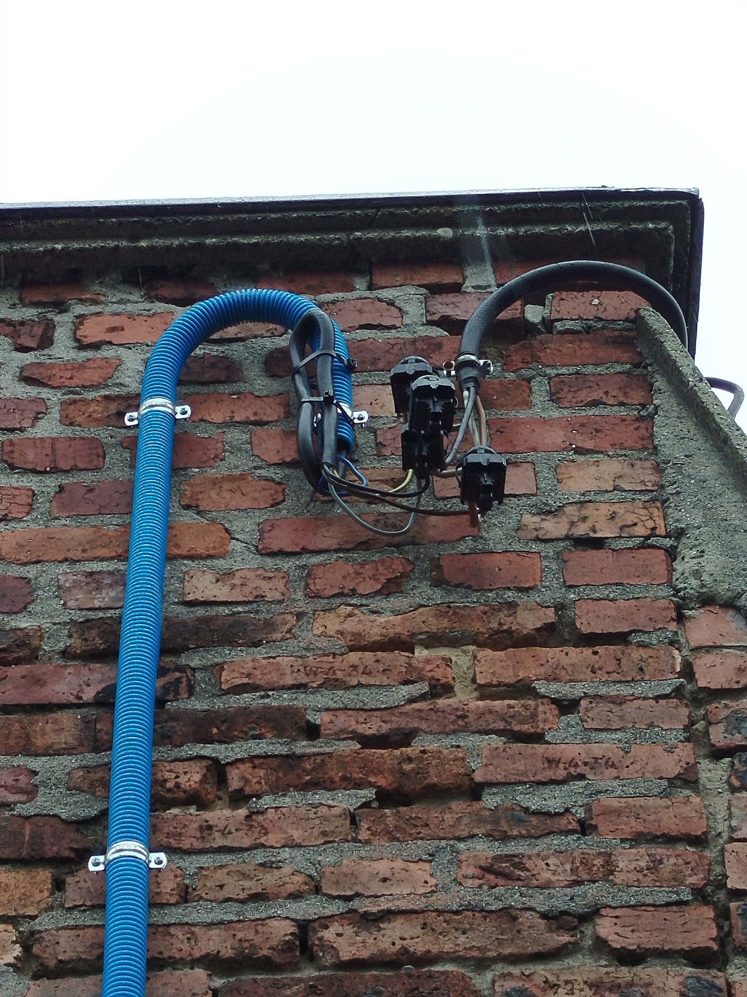Zewnętrzna instalacja elektryczna na ceglanej ścianie budynku, z niebieską rurą karbowaną i czarnymi elementami łączeniowymi, przymocowana metalowymi uchwytami.