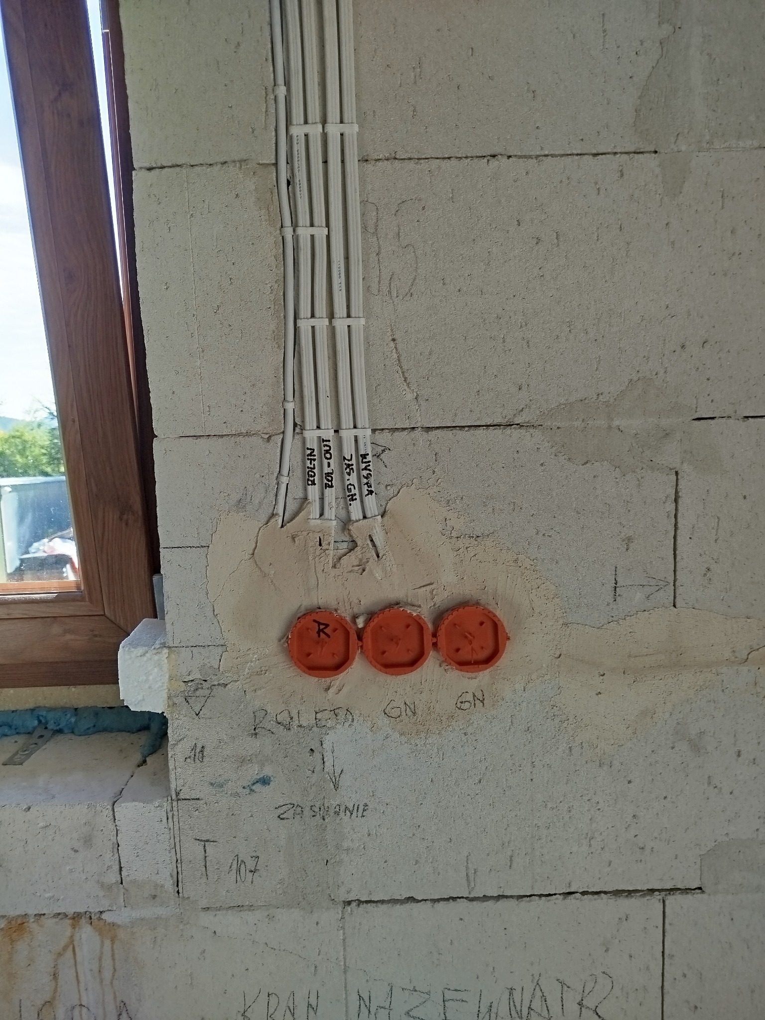 Przygotowanie do rozprowadzenia instalacji elektrycznej w surowym budynku: widoczne kable w peszlach, puszki instalacyjne w ścianie z betonu komórkowego oraz odręczne notatki na ścianie.