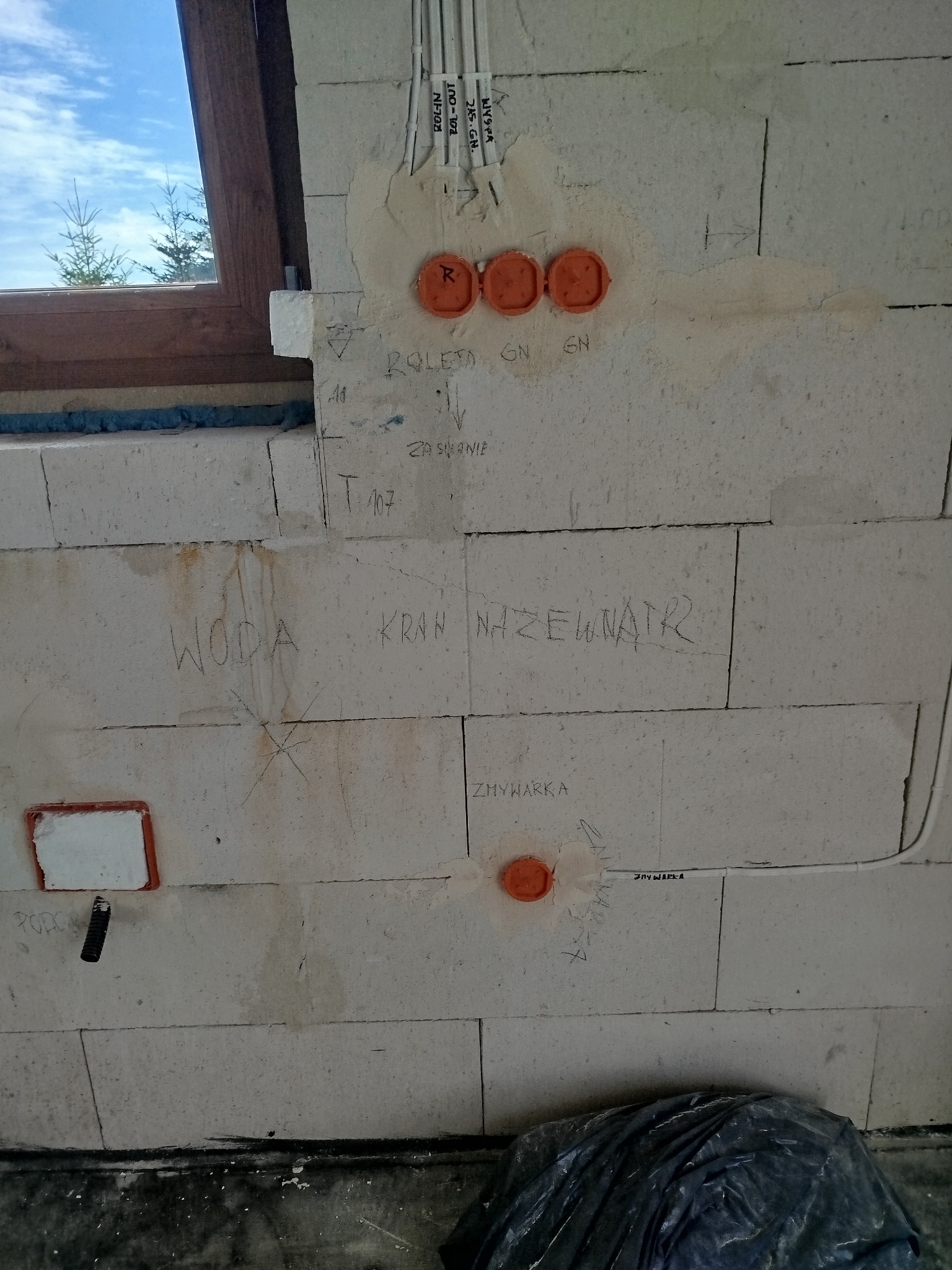 Ściana z betonu komórkowego w trakcie prac instalacyjnych, widoczne puszki elektryczne, kable w peszlach, instalacja wodna oznaczona napisem 'WODA KRAN NAZEWNATR', okno dachowe w tle.