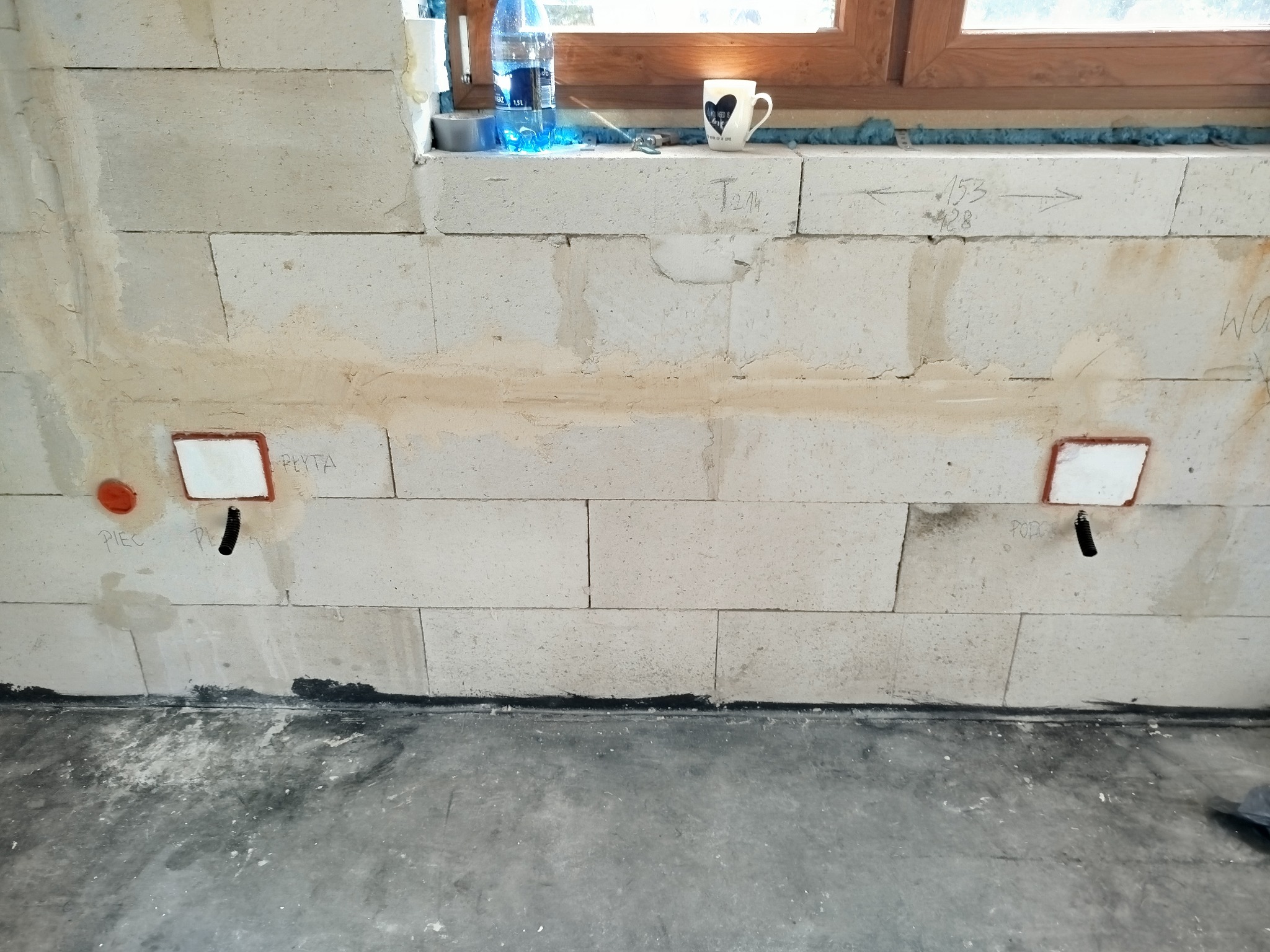 Ściana z betonu komórkowego z przygotowanymi otworami i rurami do instalacji elektrycznej, widoczne oznaczenia pomiarowe i notatki na ścianie, butelka wody i kubek na parapecie.