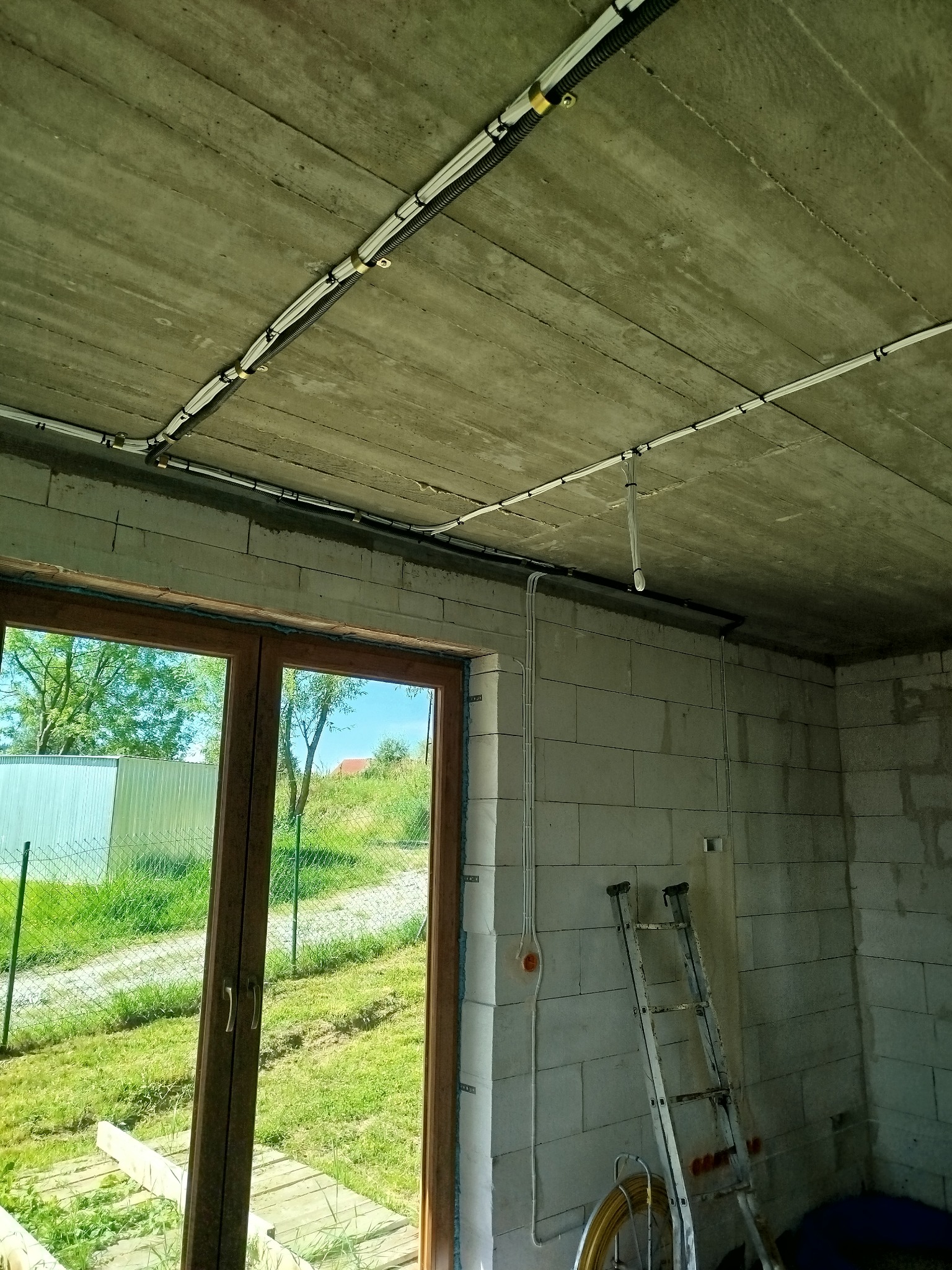 Surowy strop i ściany z betonu komórkowego w trakcie prac. Widoczne koryta kablowe i przewody elektryczne zamocowane do sufitu oraz pionowe peszle na ścianie. W tle drabina i okno z widokiem...