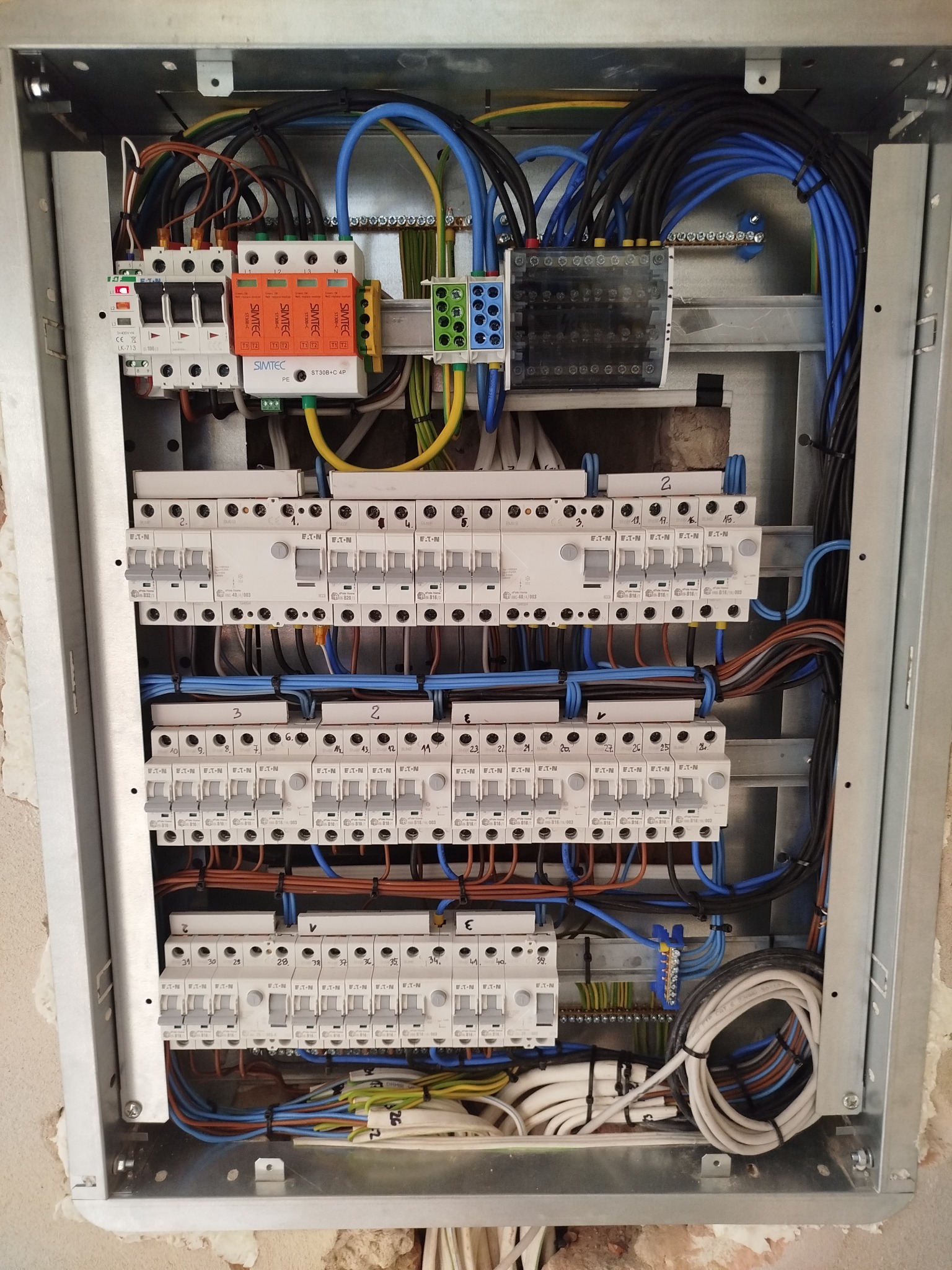 Rozdzielnica elektryczna w metalowej obudowie z widocznymi bezpiecznikami, przewodami w kolorach niebieskim, czarnym, brązowym i żółto-zielonym oraz zabezpieczeniami przeciwprzepięciowymi SIMTEC...