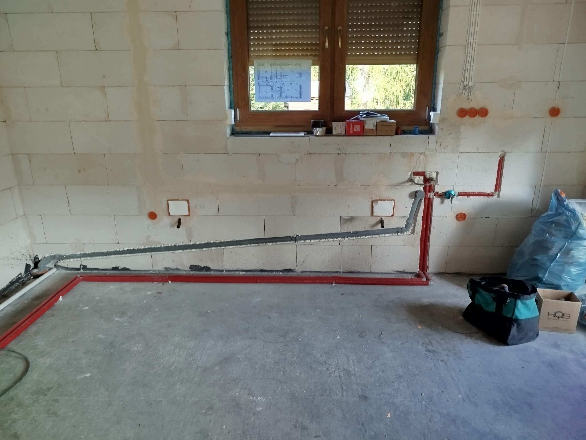 Wnętrze w trakcie remontu z widocznymi przewodami elektrycznymi i rurami w ścianach z betonu komórkowego, skrzynka narzędziowa i niebieska folia zabezpieczająca w rogu pomieszczenia.