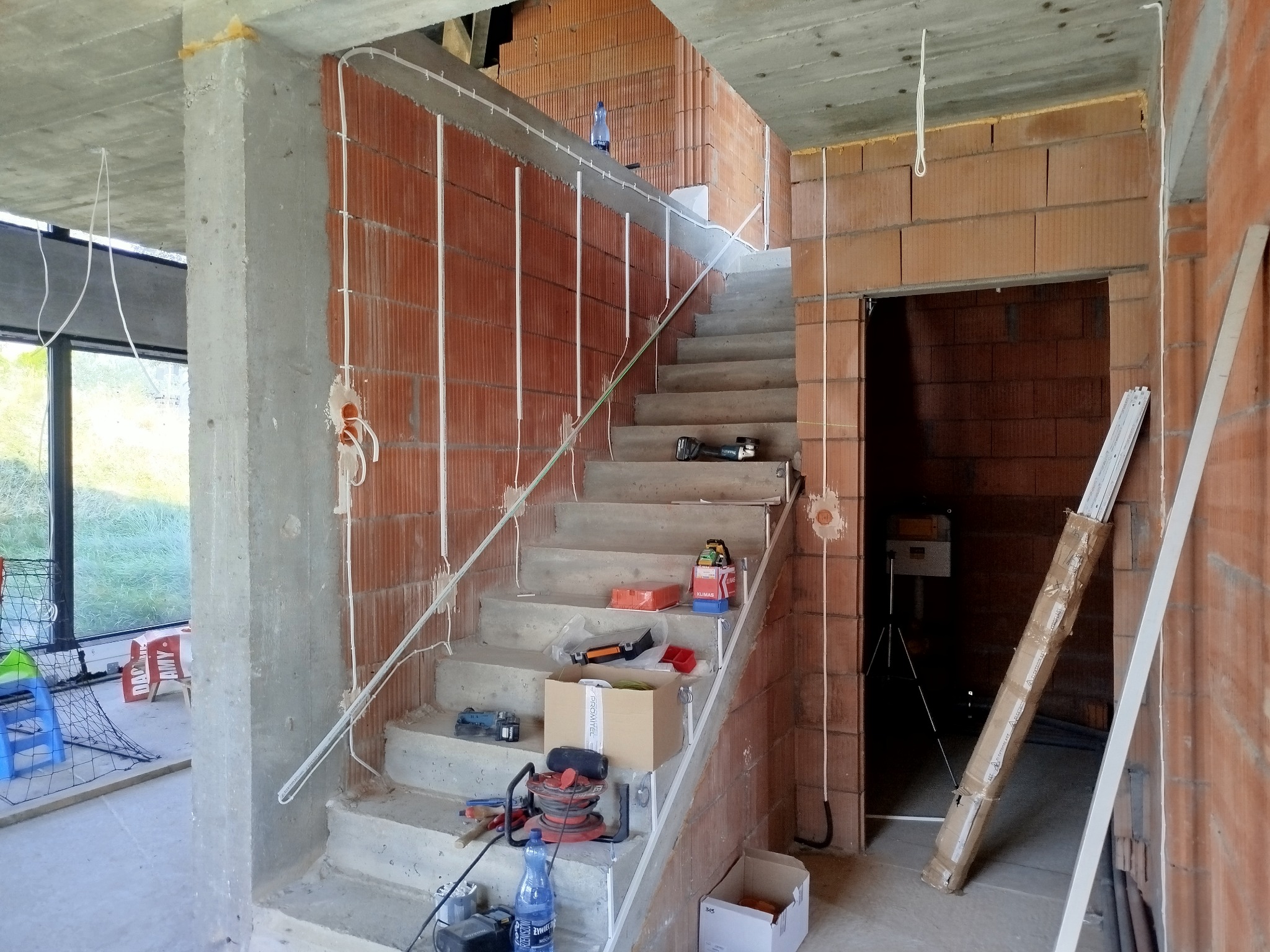 Wnętrze w trakcie remontu: surowe schody betonowe z narzędziami, ceglana ściana z poprowadzonymi przewodami elektrycznymi w białych listwach, widoczny otwór drzwiowy i kolumna betonowa.