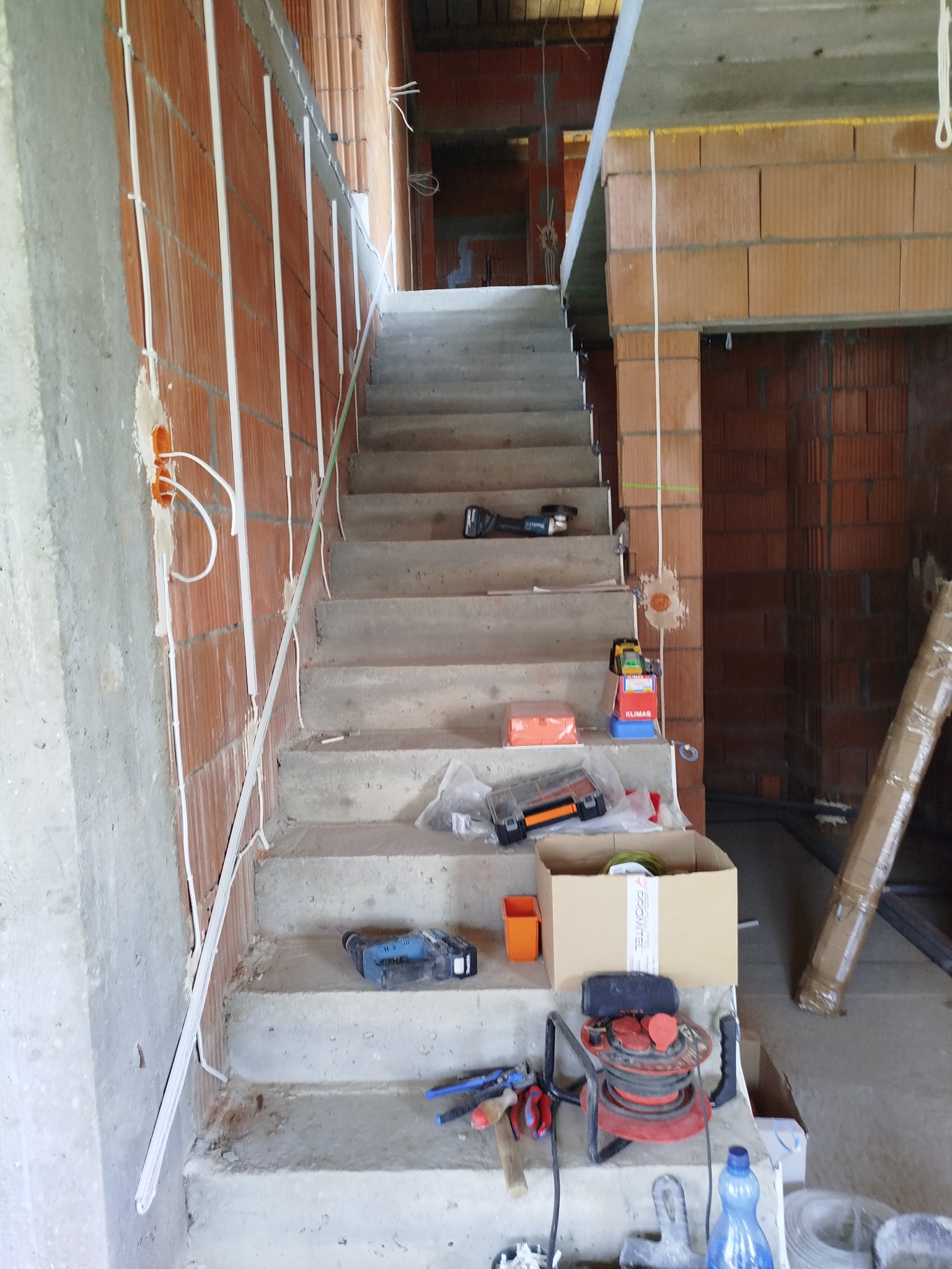 Niedokończone betonowe schody w surowym stanie z widocznymi przewodami elektrycznymi przymocowanymi do ściany z cegieł, narzędzia i materiały budowlane porozrzucane na stopniach.