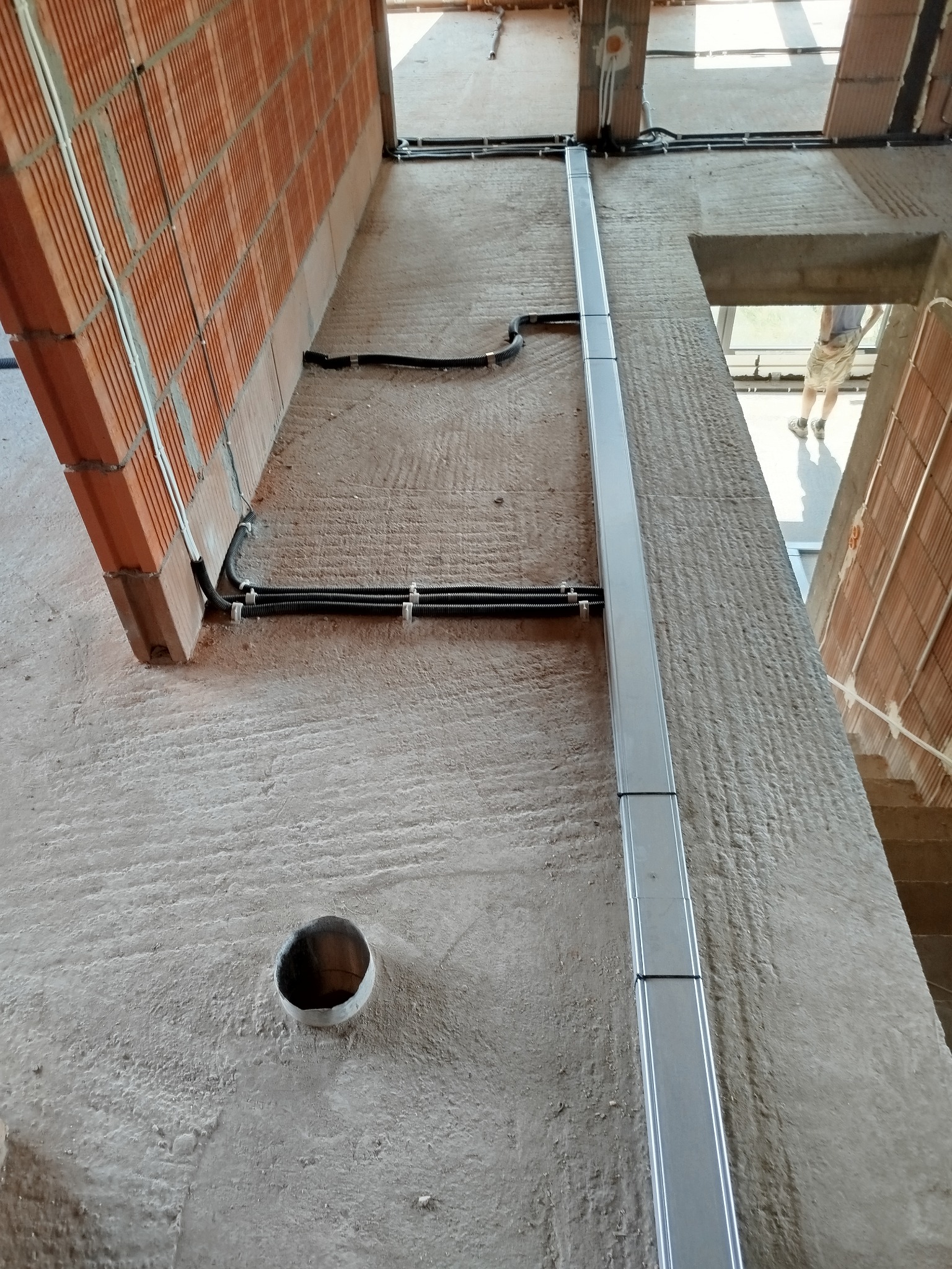 Wewnętrzna instalacja elektryczna w trakcie montażu: kable w peszlach mocowane do ściany z cegły, pionowa metalowa listwa maskująca, widoczny otwór w ścianie. Surowy stan wykończenia wnętrza.