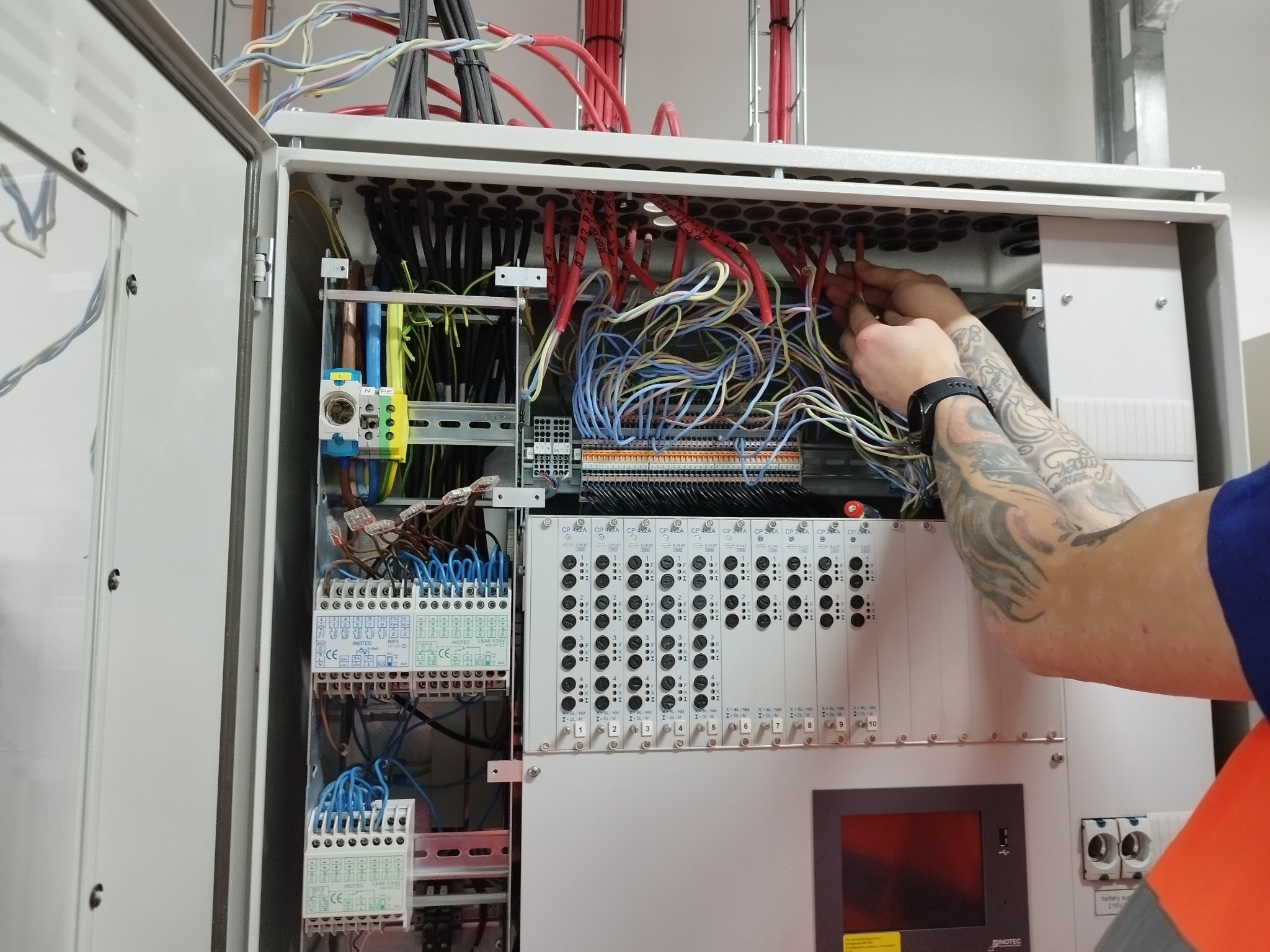 Otwarta szafa sterownicza z licznymi przewodami i elementami elektrycznymi, ręce elektryka z tatuażem podłączają czerwone kable w górnej części panelu.